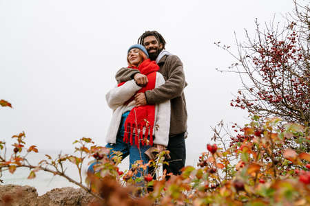 Lovely young multiethnic couple cuddling at the beach, wearing warm clothesの写真素材