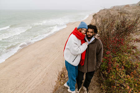 Lovely young multiethnic couple cuddling at the beach, wearing warm clothesの写真素材