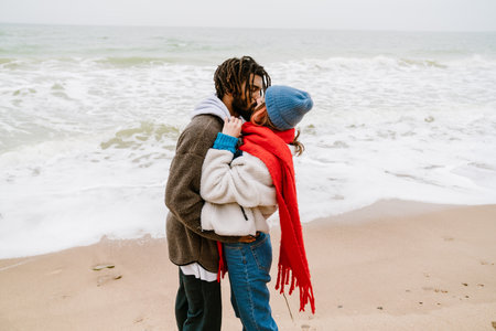 Lovely young multiethnic couple cuddling at the beach, wearing warm clothesの写真素材