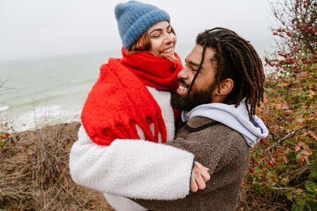 Lovely young multiethnic couple cuddling at the beach, wearing warm clothesの写真素材