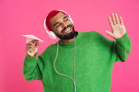 Joyful african american guy in headphones holding paper plane isolated over pink background