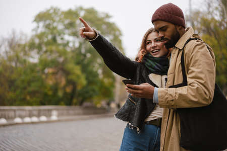 Multinational beautiful couple pointing finger aside and using mobile phone at city street
