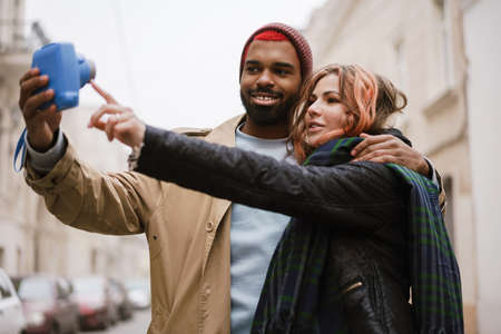 Multinational smiling couple hugging and taking selfie on retro camera at city street