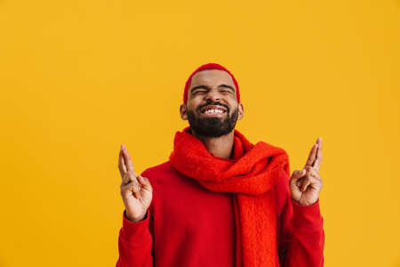 Excited african american guy holding fingers crossed for good luck isolated over yellow background