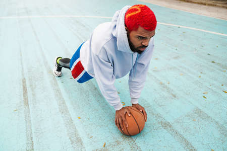 Bearded african american guy in earphones doing exercise with basketball at playground outdoorsの写真素材