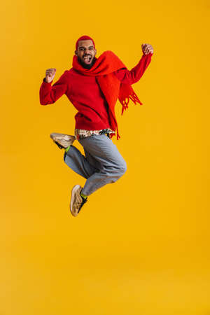 Excited happy african man wearing scarf having fun isolated over yellow background, full length, jumpingの写真素材