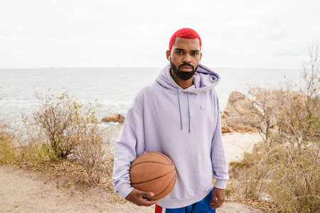 Bearded african american guy in earphones standing with basketball on beach