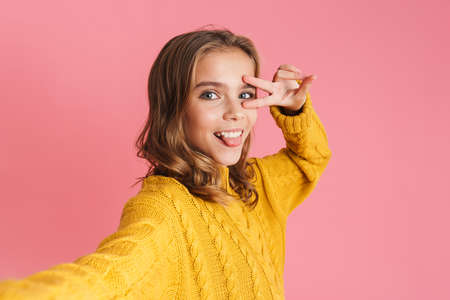 Excited beautiful girl gesturing peace sign while taking selfie photo isolated over pink background