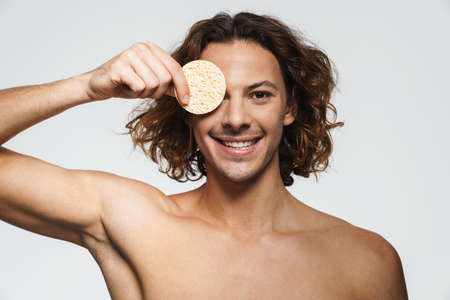 Happy shirtless guy smiling while posing with cosmetic sponge isolated over white backgroundの写真素材
