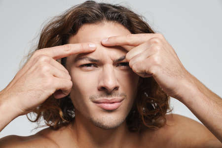 Displeased shirtless guy squeezing pimple on his face isolated over white background