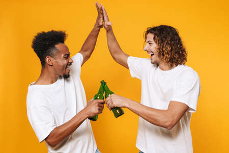 Portrait of a two cheerful young men best friends toasting with beer bottles isolated over yellow backgroundの写真素材