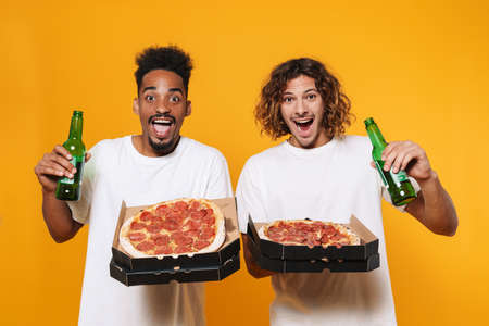 Portrait of a two satisfied young men watching football while drinking beer and eating pizza isolated over yellow backgroundの写真素材
