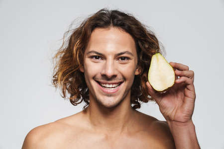 Joyful shirtless guy smiling while posing with pear isolated over white background