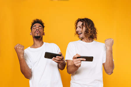 Two excited african man friends standing isolated over yellow background, playing games on mobile phones, celebratingの写真素材