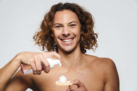Laughing shirtless guy using cosmetic sponge and cleansing foam isolated over white backgroundの写真素材