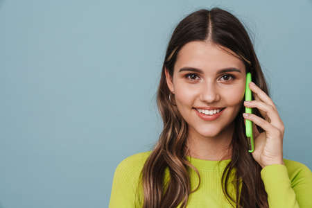 Joyful beautiful cute girl smiling while talking mobile phone isolated over blue background