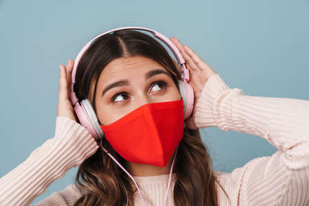 Caucasian nice girl in face mask listening music with headphones isolated over blue backgroundの写真素材