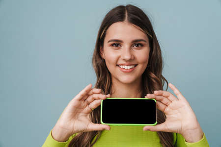 Happy beautiful cute girl smiling and showing mobile phone isolated over blue background