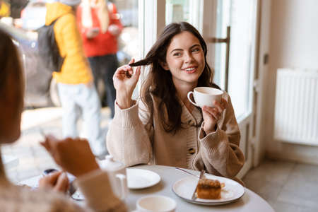 Two lovely smiling young women friends having lunch while sitting in cafe indoors, drinking teaの写真素材