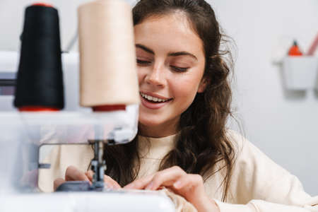 Nice happy girl seamstress smiling while working with sewing machine in atelierの写真素材
