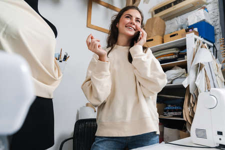 Smiling nice brunette girl seamstress talking on mobile phone while working in atelierの写真素材