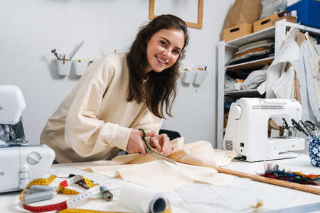 Happy nice brunette girl seamstress cutting fabric while working in atelierの写真素材