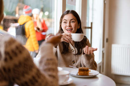 Two lovely smiling young women friends having lunch while sitting in cafe indoors, drinking teaの写真素材
