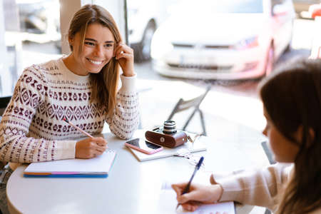 Happy charming two students girls doing homework together while sitting in cafe indoorsの写真素材