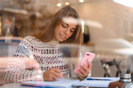 Pleased charming student girl using mobile phone while doing homework in cafe indoors