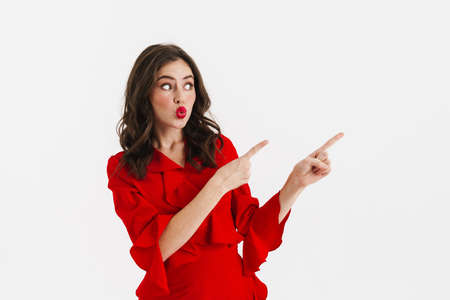 Surprised nice girl wearing red dress pointing fingers aside isolated over white backgroundの写真素材