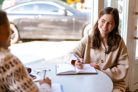 Happy charming two students girls doing homework together while sitting in cafe indoorsの写真素材