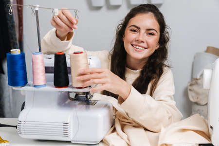 Nice happy girl seamstress smiling while working with sewing machine in atelier