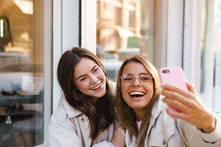 Two cheerful girls friends taking selfies while sitting in cafe outdoorsの写真素材