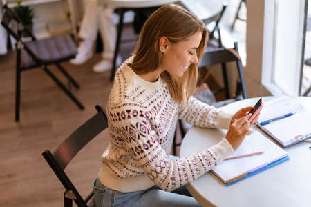 Smiling charming student girl using mobile phone while doing homework in cafe indoorsの写真素材