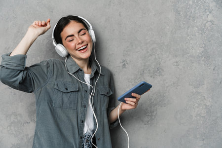 Happy attractive young woman listening to music with headphones, holding mobile phone isolated over gray background, dancingの写真素材