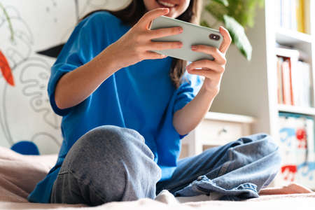 Joyful brunette girl playing online game on mobile phone while sitting on bed at home