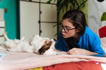 Happy girl in eyeglasses stroking her lovely dog while lying on bed at homeの写真素材