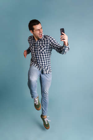Happy young casual man taking a selfie while jumping isolated over blue background, full length