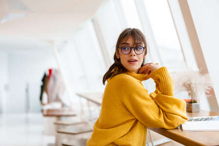 Charming pleased nice girl in eyeglasses looking aside while reading book in cafe indoors