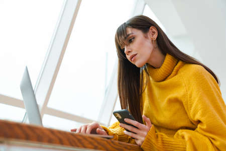 Charming serious girl using cellphone and working with laptop while sitting at table indoors