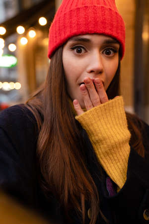 Charming shocked girl in hat covering her mouth and taking selfie photo at city street