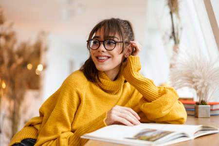 Charming beautiful smiling girl in eyeglasses reading book while sitting in cafe indoors