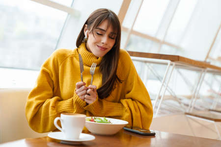 Charming happy girl in earphones smiling and using mobile phone while having lunch in cafe indoorsの写真素材