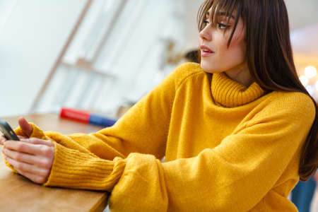 Charming focused girl using mobile phone while sitting at table indoorsの写真素材