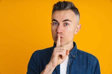 Serious handsome man showing silence gesture at camera isolated over yellow background