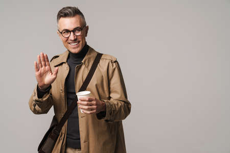 Handsome man wearing stylish coat posing isolated over gray background, carrying bag, drinking coffee, waving handの写真素材