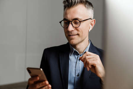 Smiling grey man in earphones working with cellphone and computer at office