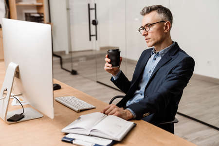 Focused grey man drinking coffee while working with computer in officeの写真素材