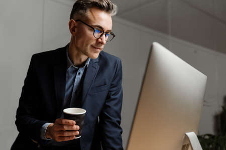 Pleased grey man drinking coffee while working with computer in office