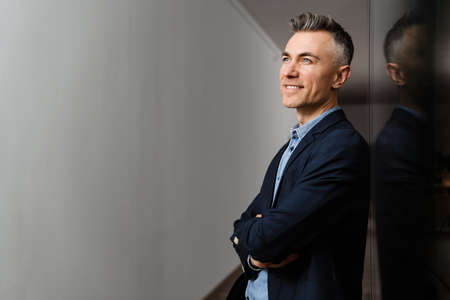 Happy grey man smiling and looking upward while leaning on wall in officeの写真素材